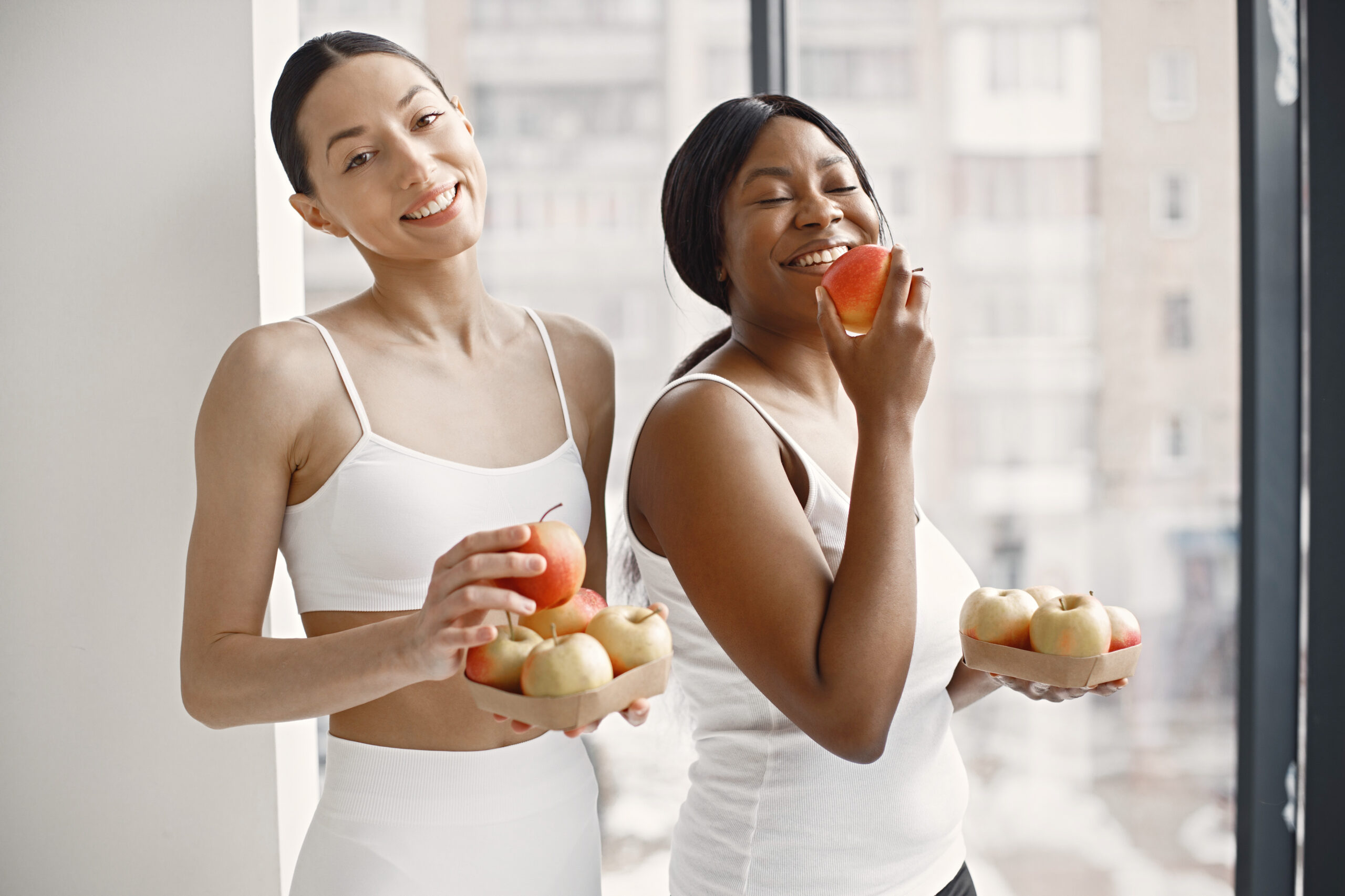 Black and caucasian women standing in studio with big windows and holding apples